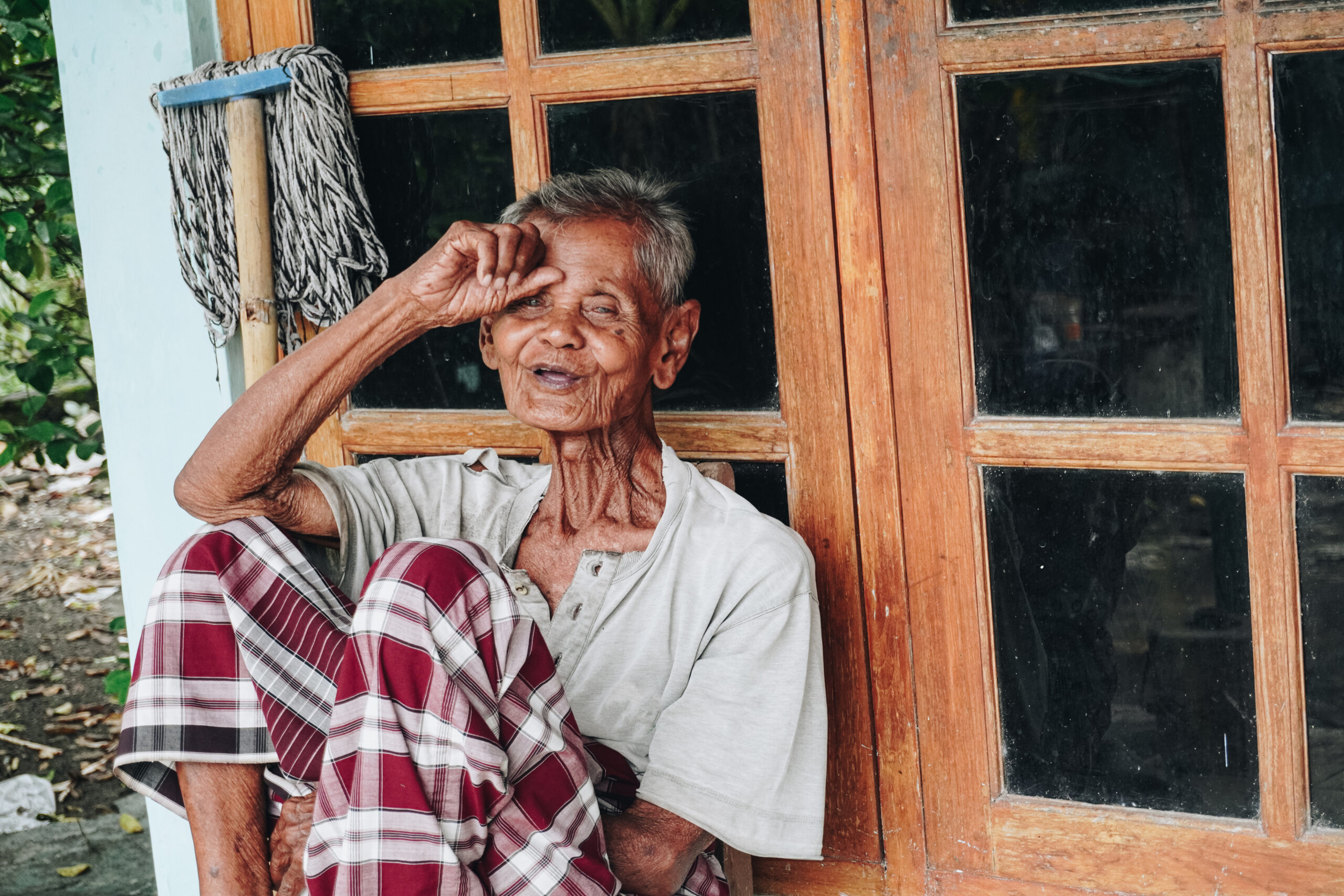 Thoughtful senior elderly man wearing saroong sitting alone in front of house with hands placed on head.